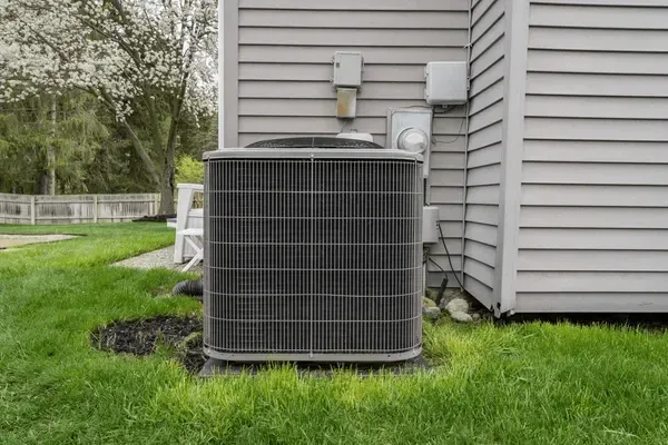 Outdoor air conditioner unit beside a gray house with a green lawn and trees in the background