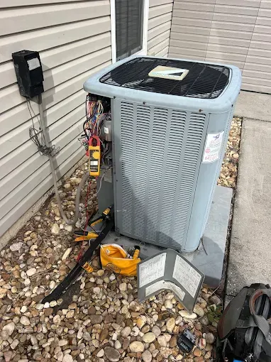 Outdoor central air conditioner unit beside a house with exposed tools and wiring on gravel.