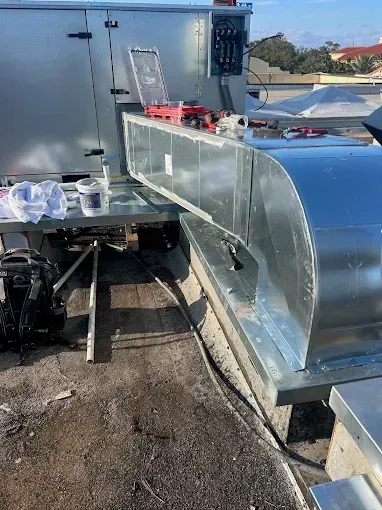Metal rooftop HVAC duct under construction beside a gray rooftop unit, with tools on top.