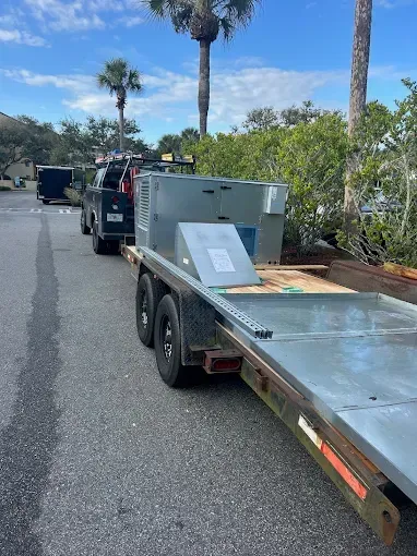 Flatbed trailer with metal equipment on a street, parked behind a truck near palm trees.