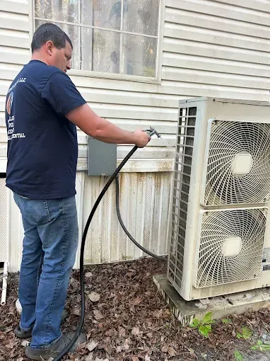 Technician cleaning an outdoor air conditioner unit with a hose beside a house.