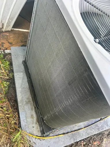 Outdoor air conditioner unit beside a building wall, with dirt and grass around the concrete pad