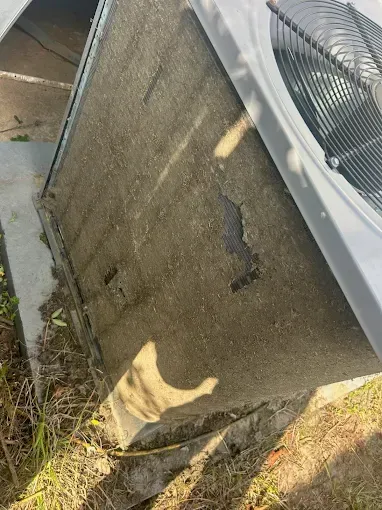 Damaged outdoor HVAC unit with cracked, stained side panel beside a concrete wall and weeds
