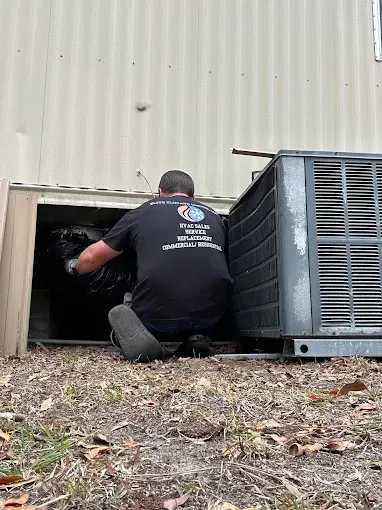 Person kneels by a wall opening, reaching into a dark crawl space beside an outdoor HVAC unit.