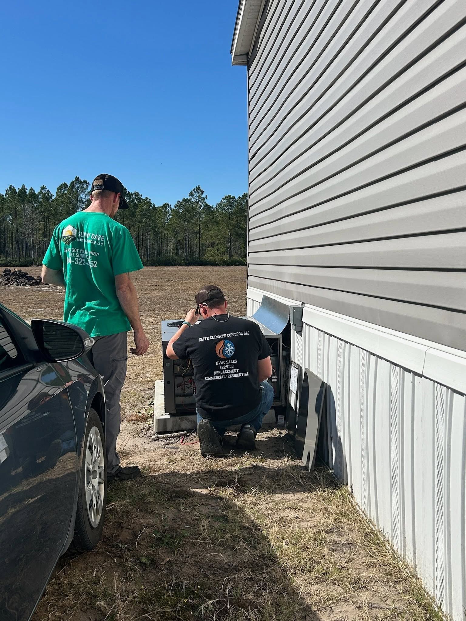 Two people working beside a house, one kneeling near the foundation and one standing by a truck.