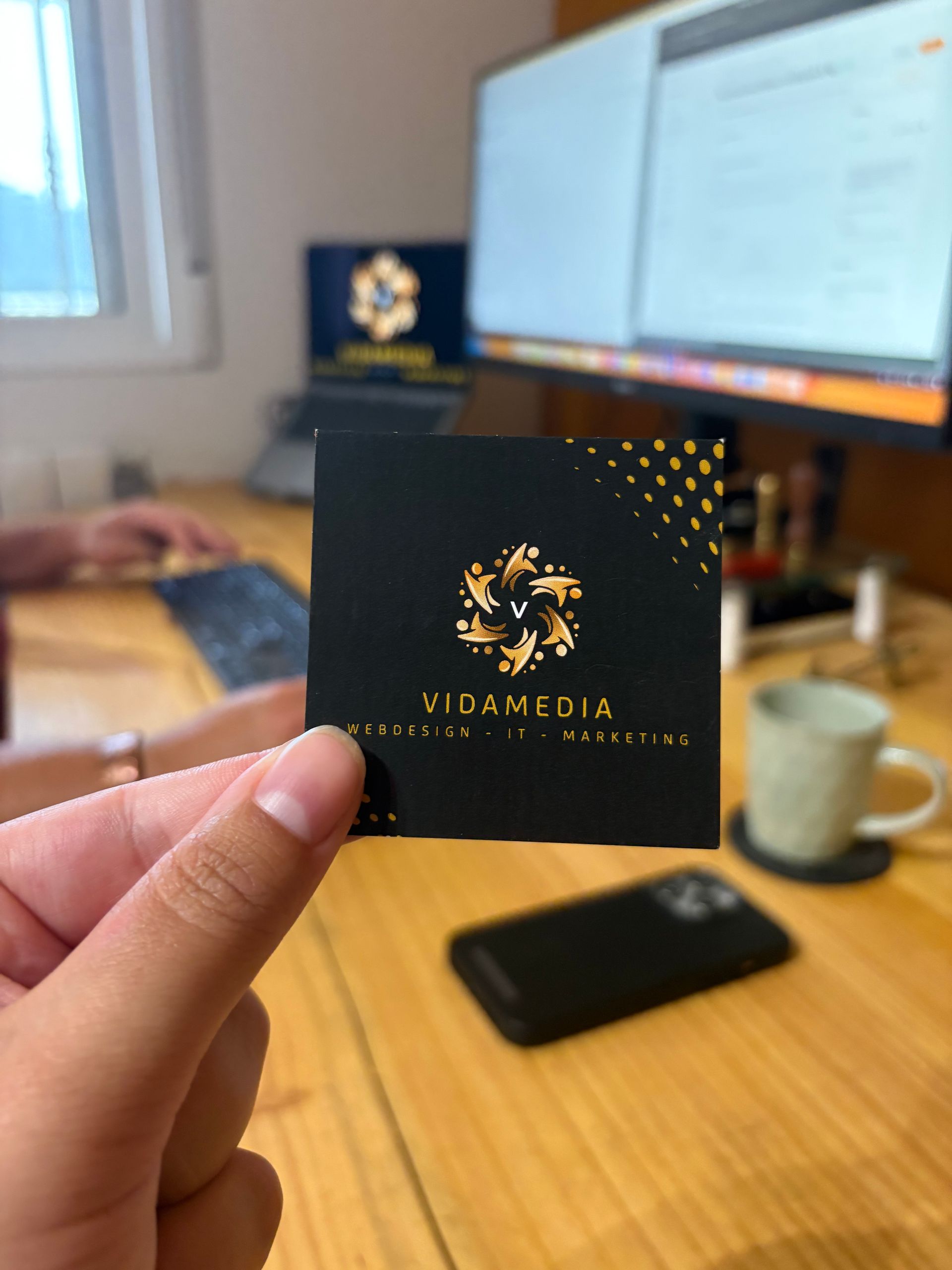 Hand holding a black business card with gold logo and text in an office setting.