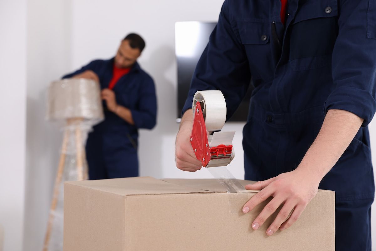 A Man is Taping a Cardboard Box With Tape — Ocean Grove Removals In Whittington, VIC