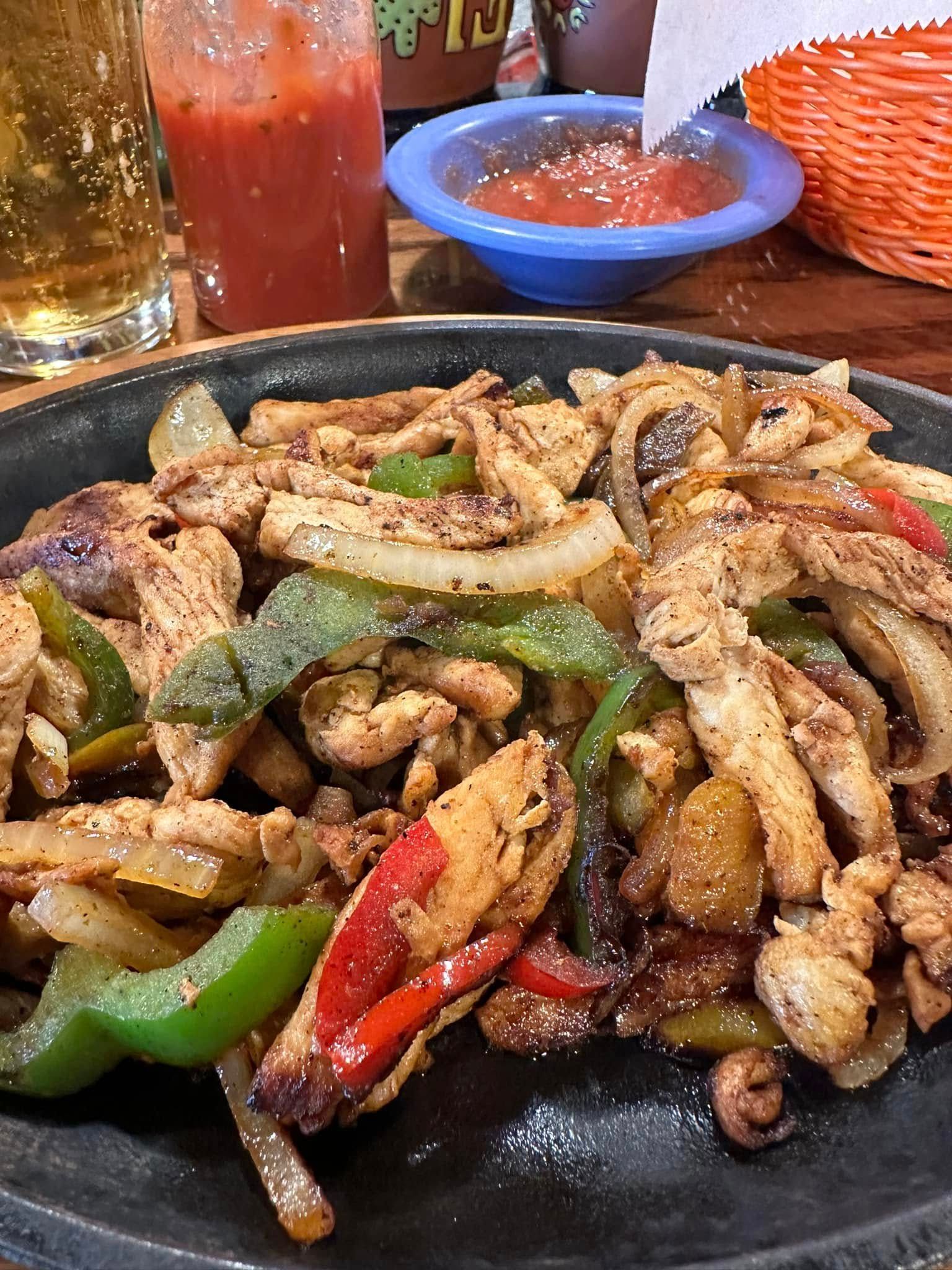 Stir-fried meat and peppers on a black plate with dipping sauce and drinks in the background