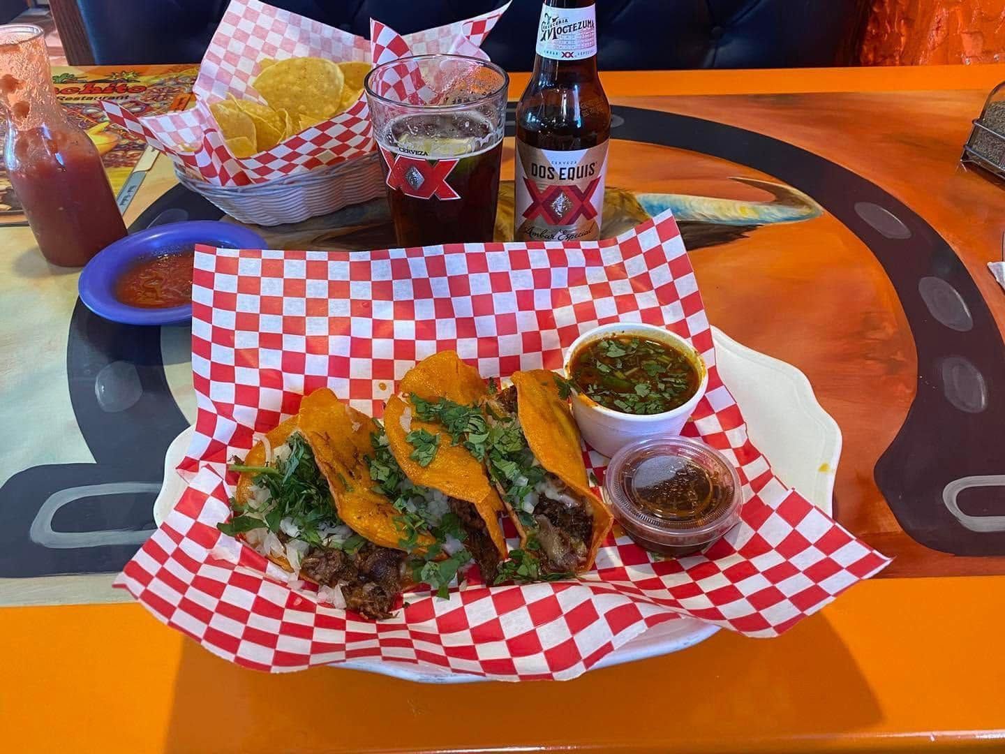 Fried tacos with green sauce and salsa on a red-checkered tray at an orange table, with beer bottles behind.