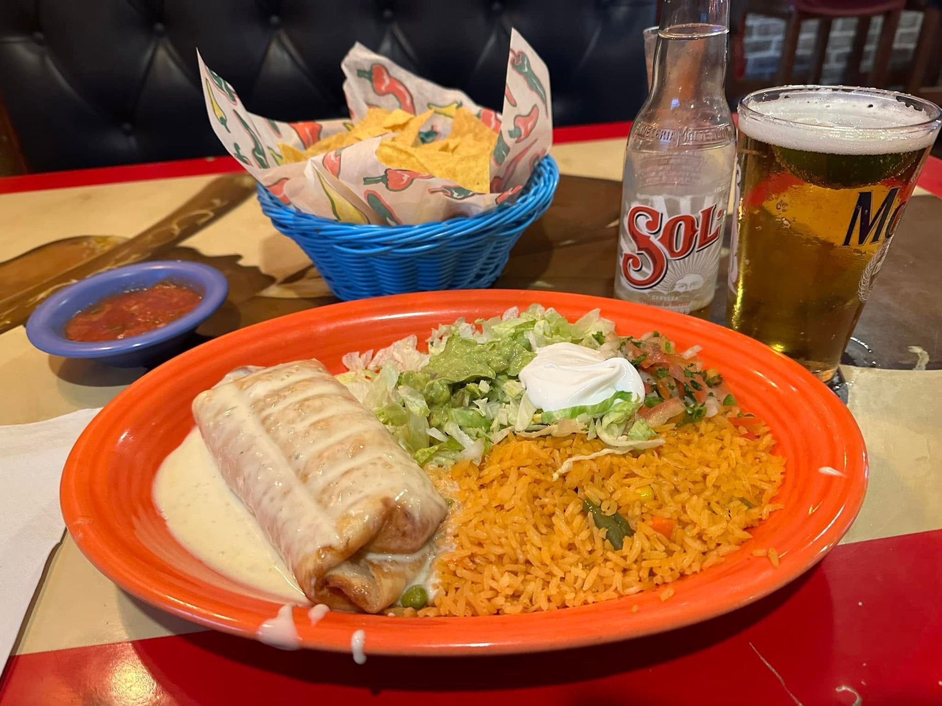 Mexican meal on orange plate with burrito, rice, lettuce, salsa, beer, and soda on a table