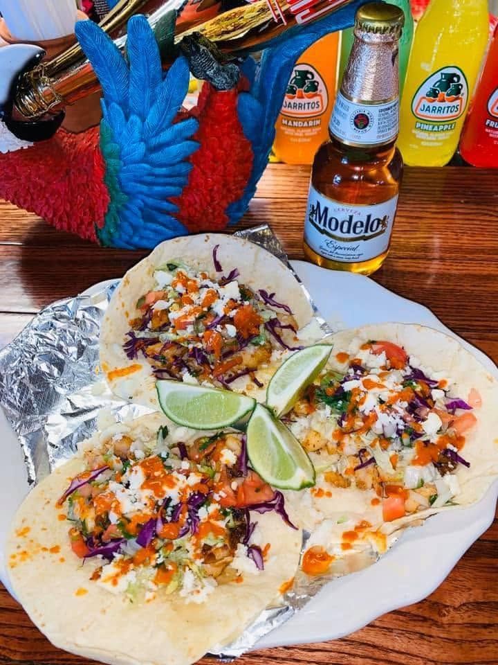 Three tacos on foil with lime wedges and a Modelo beer bottle on a wooden table.