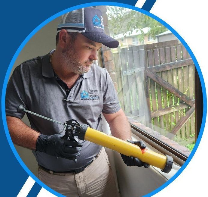A Man Wearing a Hat and Gloves is Holding a Yellow Tool in Front of a Window — Pioneer Pest Mackay In Glenella, QLD