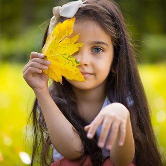 Girl with long brown hair holds a yellow leaf, covering one eye, in a sunny field.