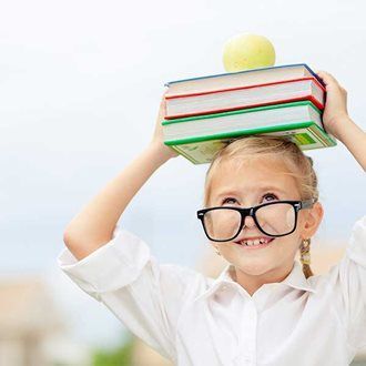 Girl wearing glasses balances books and an apple on her head, smiling.