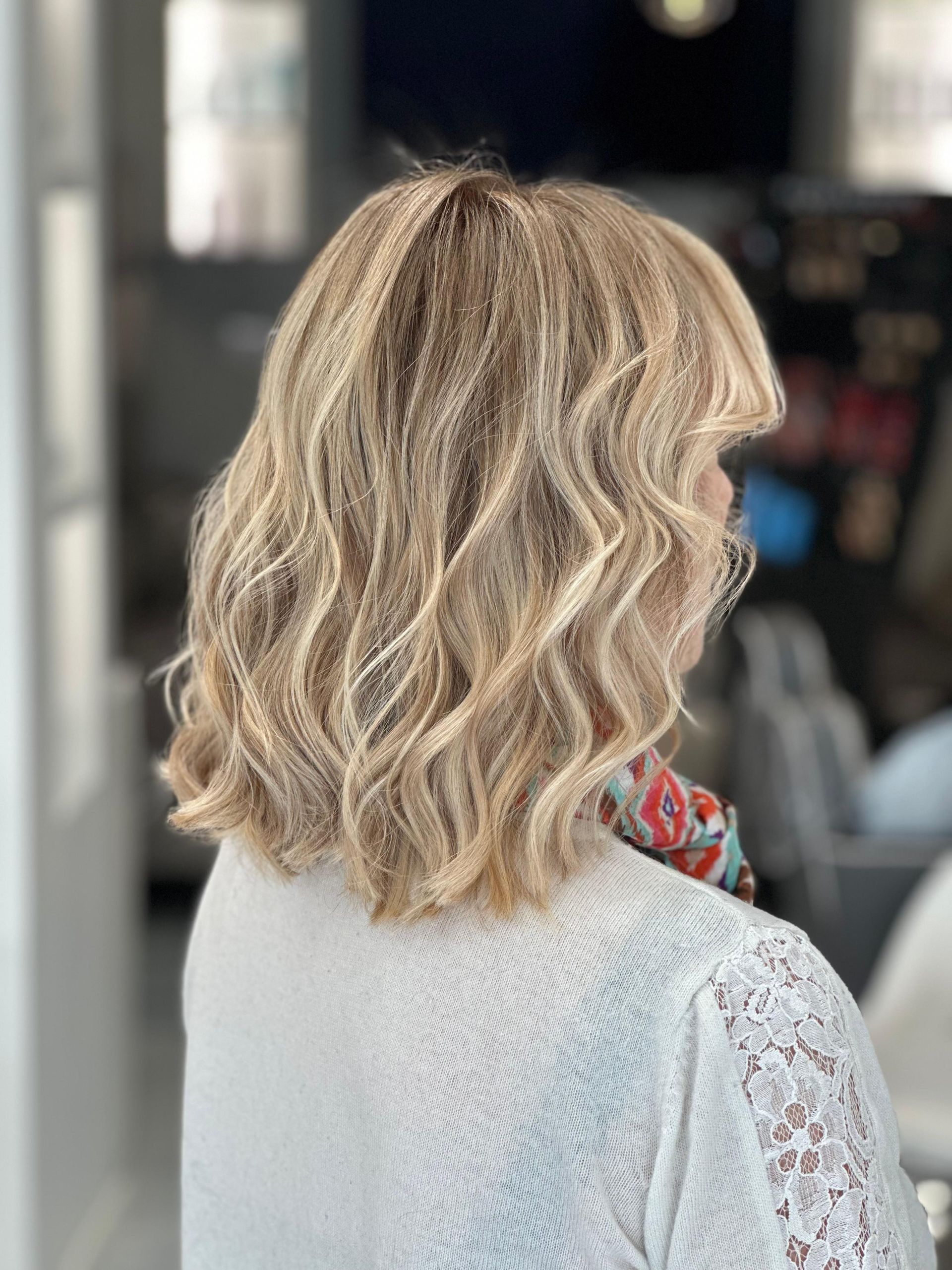 Donna con capelli biondi ondulati lunghi fino alle spalle, che indossa un maglione bianco, in un salone di bellezza.