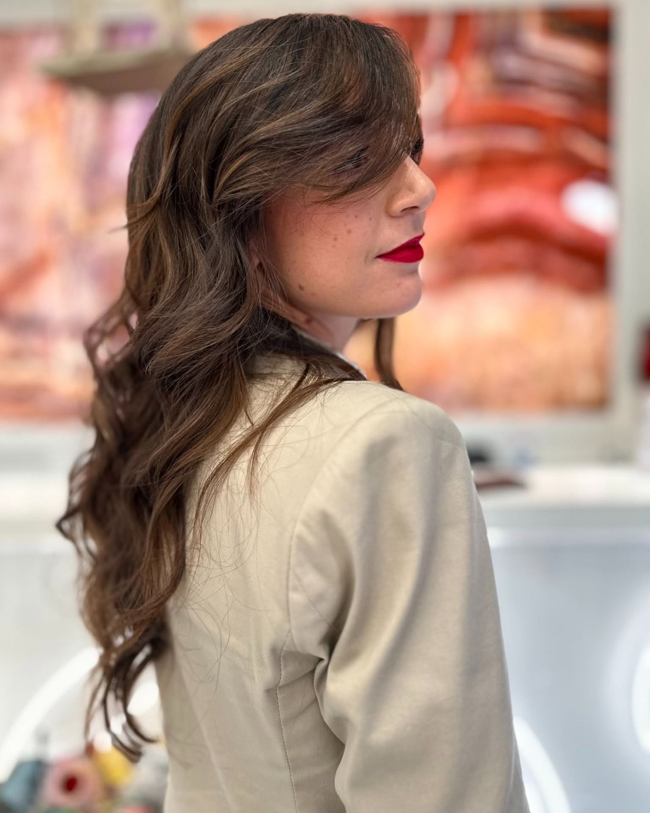 Donna con blazer beige, capelli scuri e ondulati, rossetto rosso, che guarda oltre la spalla.