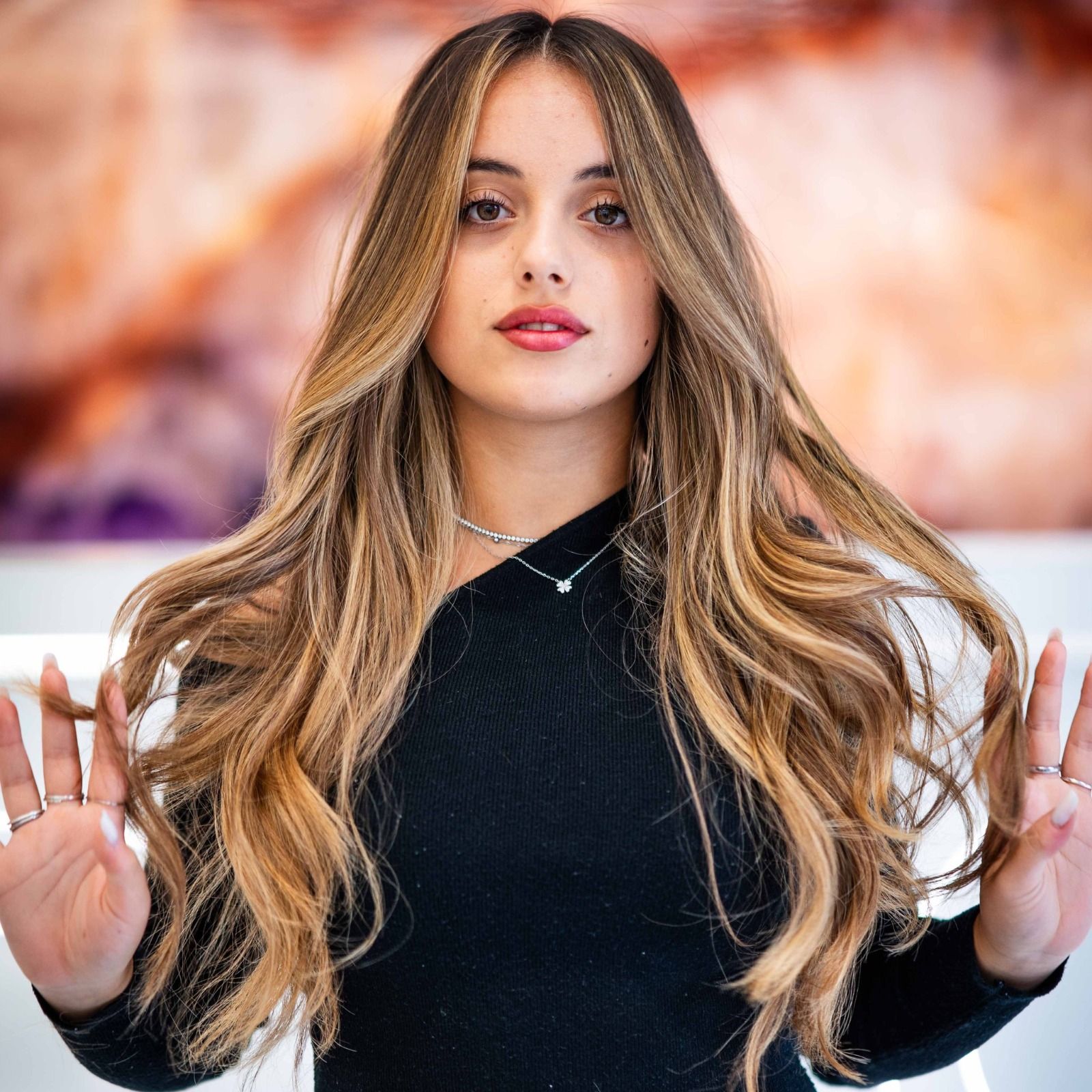 Donna con lunghi capelli biondi e castani ondulati, che indossa un top nero, posa con le mani alzate.