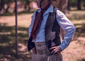 Persona con camisa azul, chaleco y bufanda, de pie al aire libre con las manos en las caderas.