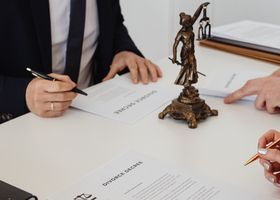 Manos firmando documentos alrededor de una estatua de bronce de la Dama de la Justicia
