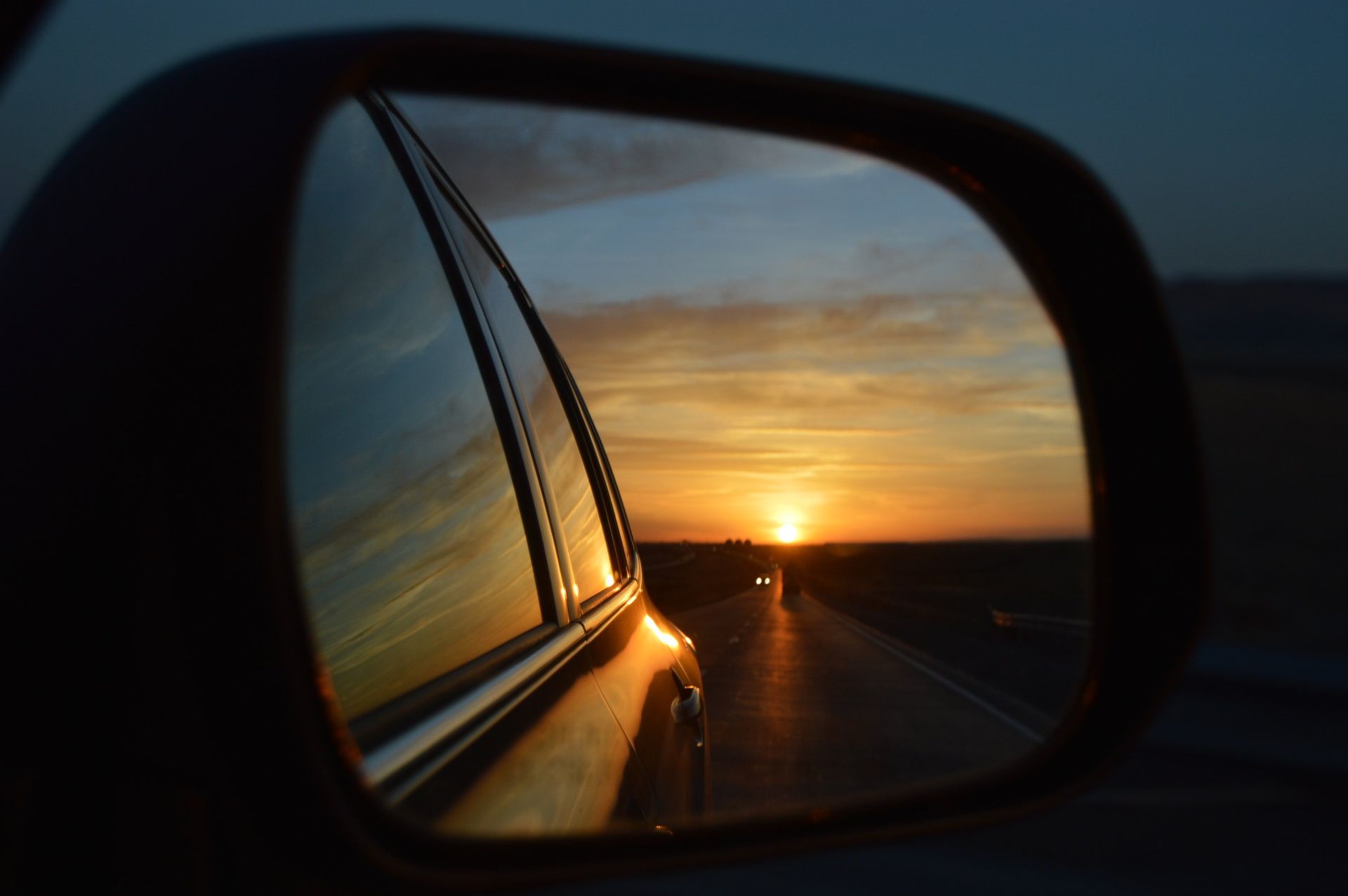 De zonsondergang wordt weerspiegeld in de zijspiegel van een auto, waardoor een weg zichtbaar is die zich uitstrekt.
