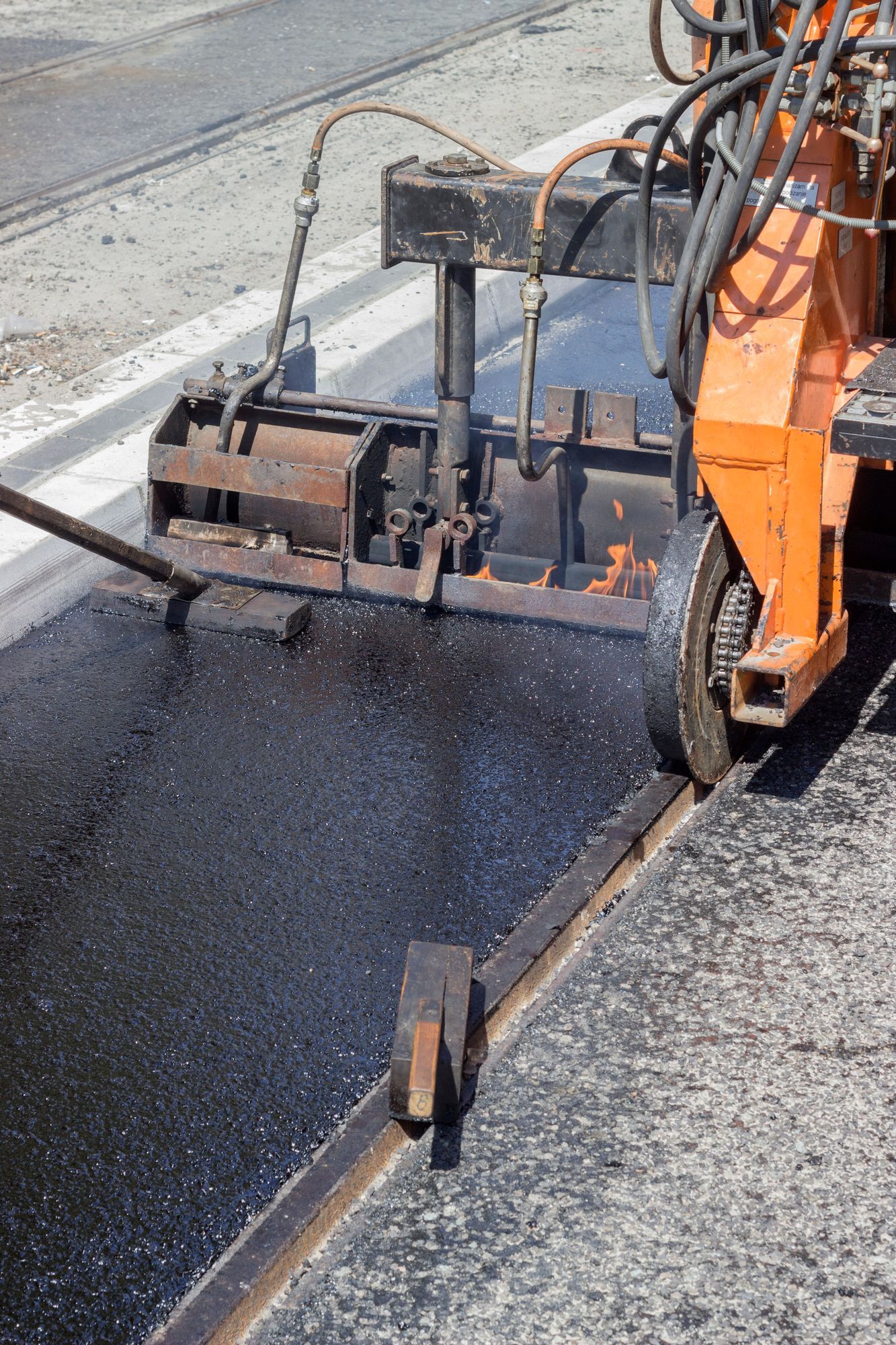 Un veicolo da cantiere arancione sta asfaltando una strada con asfalto fresco e scuro lungo un cordolo di cemento.