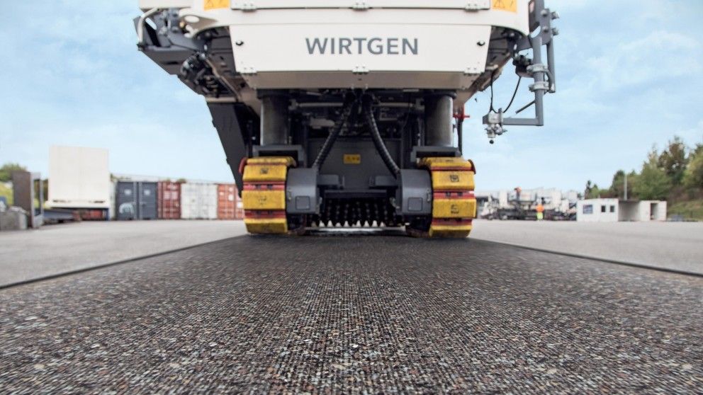 Una fresatrice a freddo Wirtgen vista dal basso, che mostra i cingoli e la superficie di asfalto appena texturizzata sottostante.