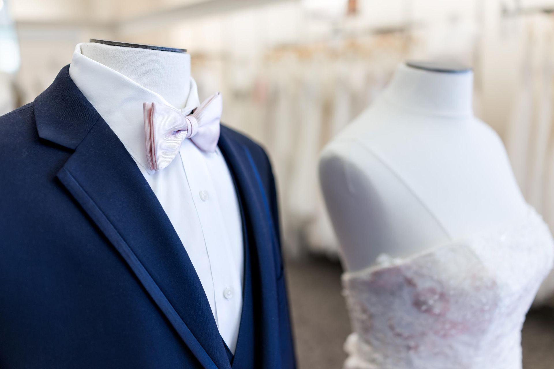 Navy suit with pink bow tie on a mannequin next to a wedding dress mannequin.