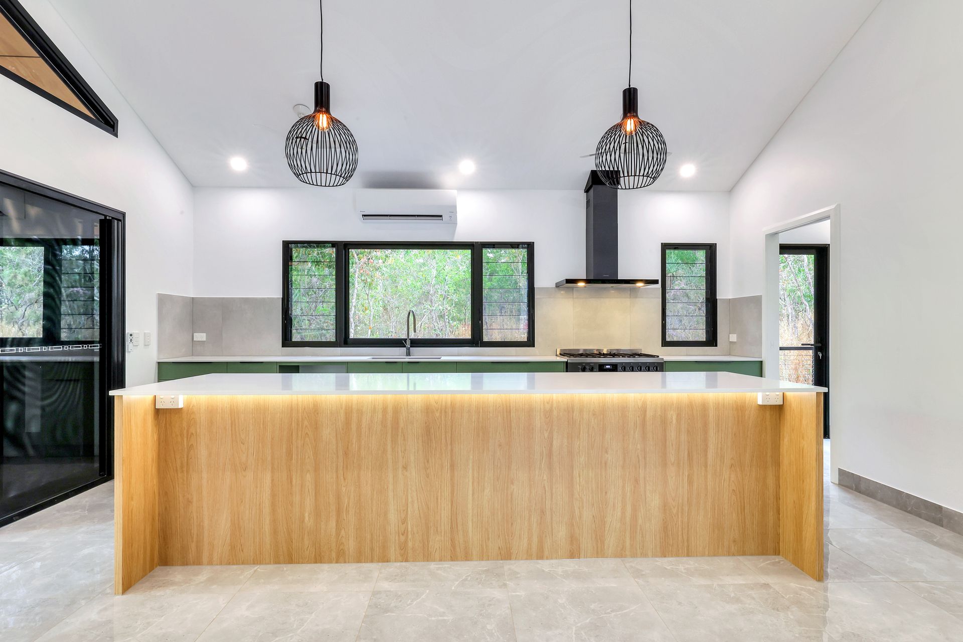 Modern kitchen with wooden island, black range hood, and pendant lights. Green countertops and windows overlooking nature — RMB Air Conditioning & Electrical in Winnellie, NT