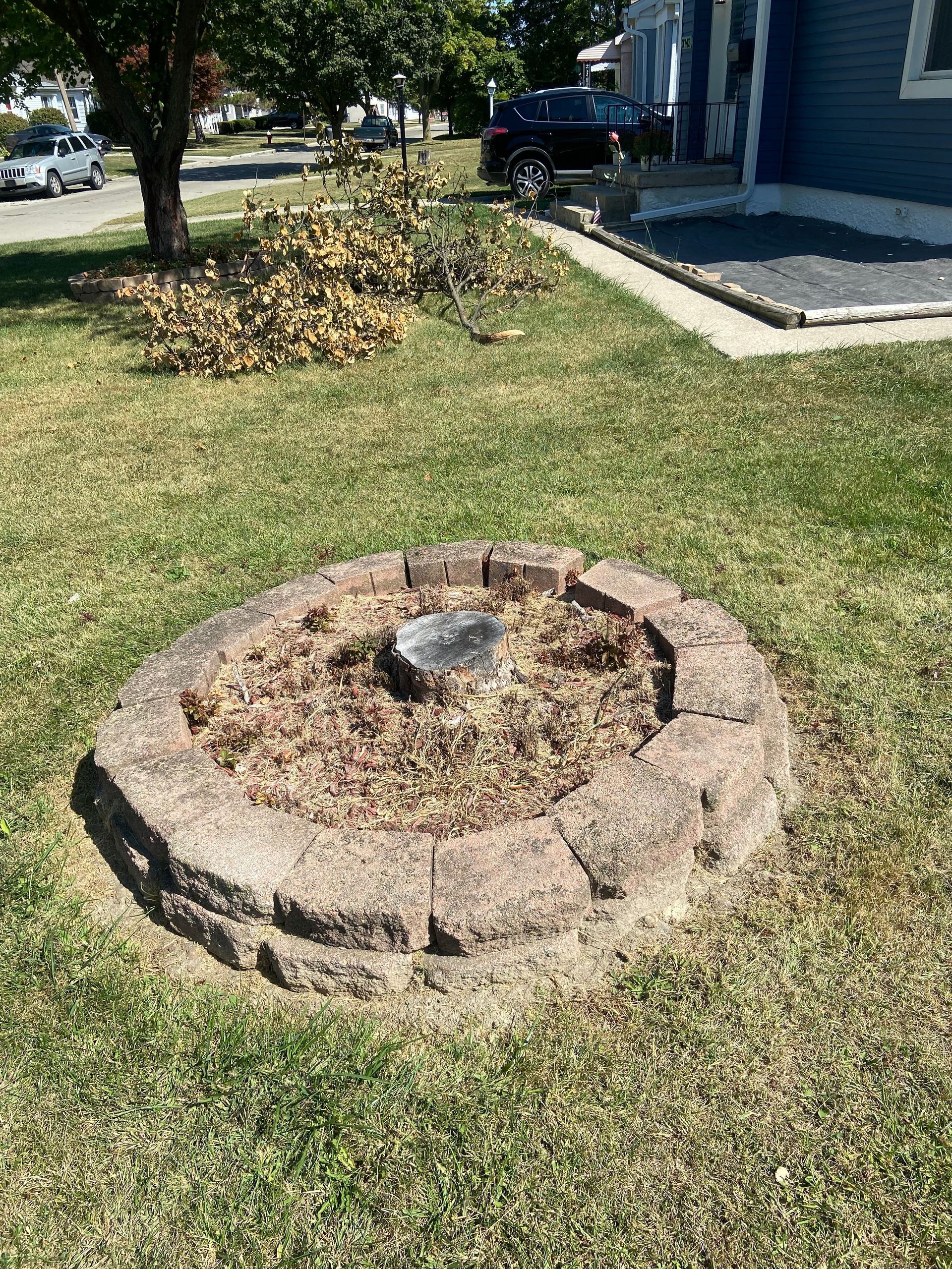 Circular stone fire pit in a grassy yard beside a house, with a pile of leaves in the background