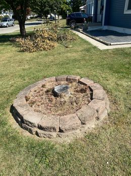 Circular stone fire pit in a grassy yard beside a house, with a pile of leaves in the background