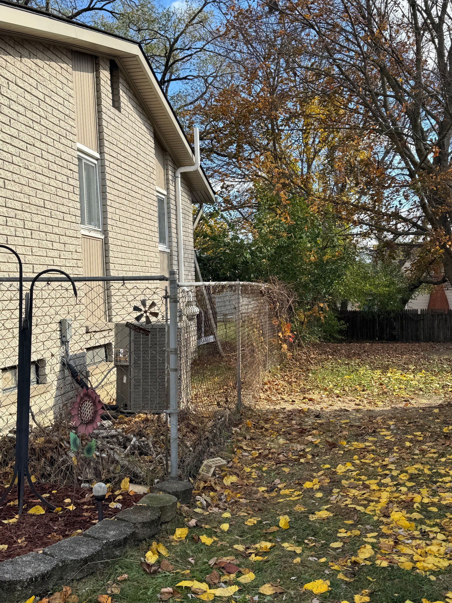 Backyard beside a tan house, with a fenced HVAC unit and autumn leaves scattered on the ground.