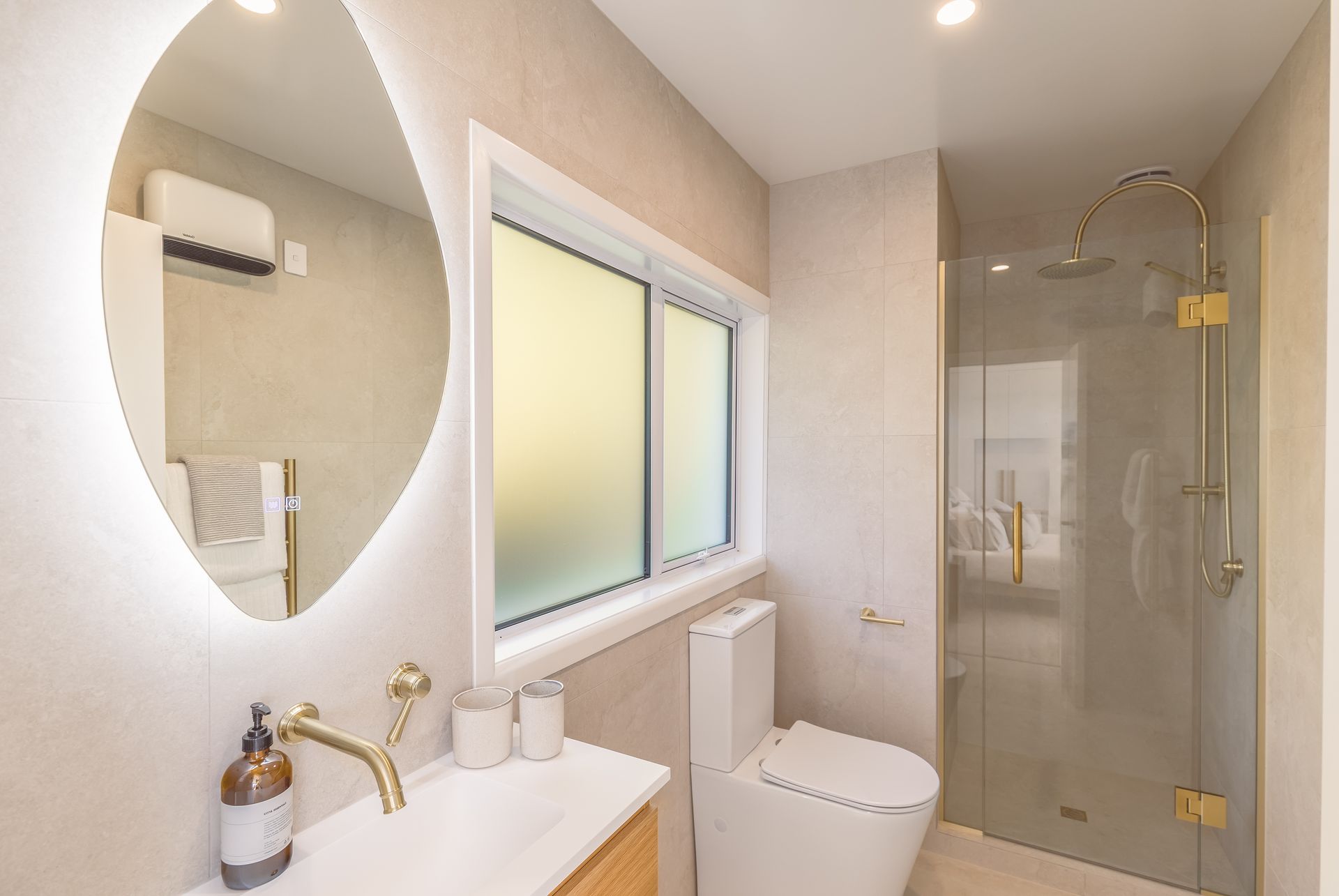 Modern bathroom with oval mirror, vanity sink, toilet, frosted window, and glass shower door.