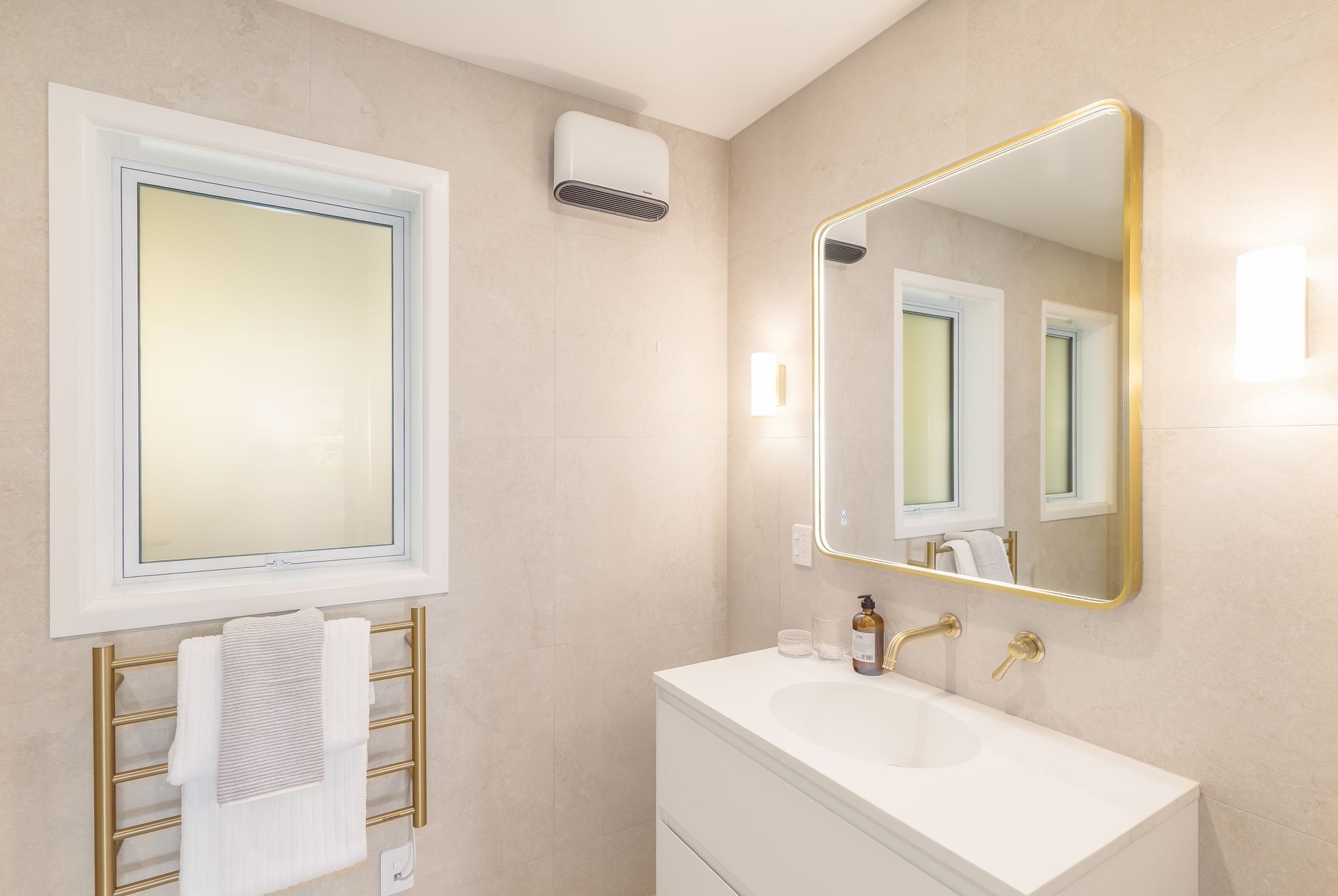 Bright bathroom with a white sink, gold-framed mirror, towel rack, and frosted window.