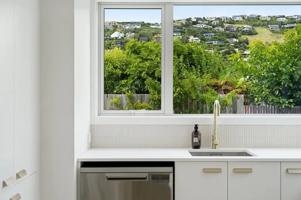 Bright kitchen with white cabinets, sink, and a large window overlooking green trees.