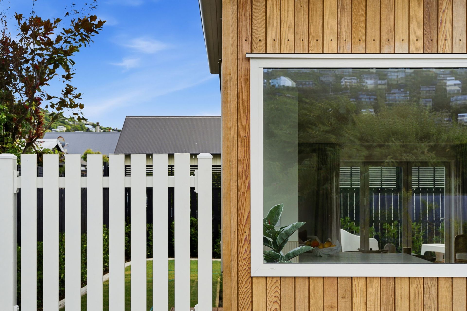 White picket fence beside a wooden house with a large window and trees reflected in the glass