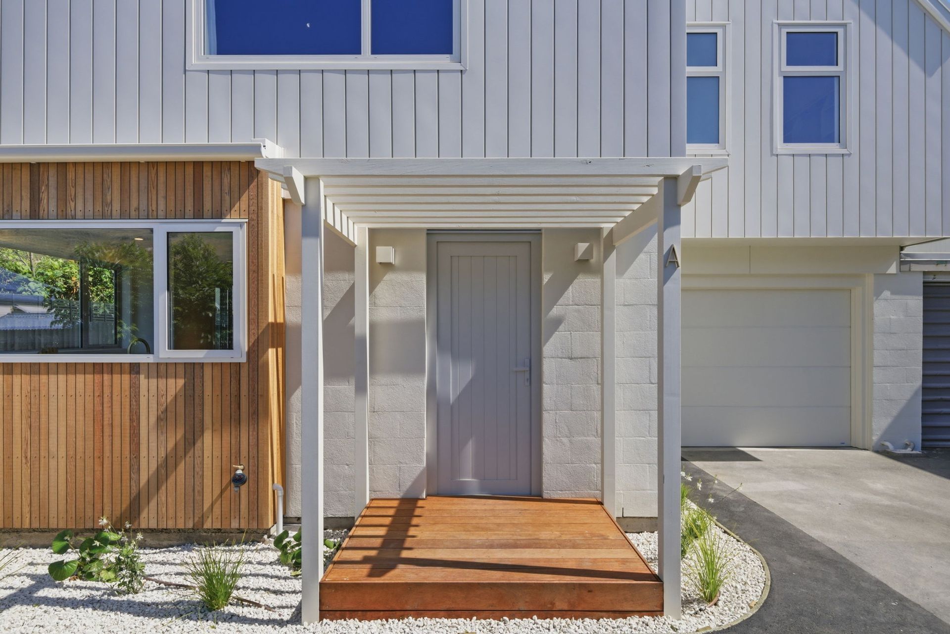 Front entrance of a modern townhouse with white siding, gray door, wooden porch, and driveway garage on the right.