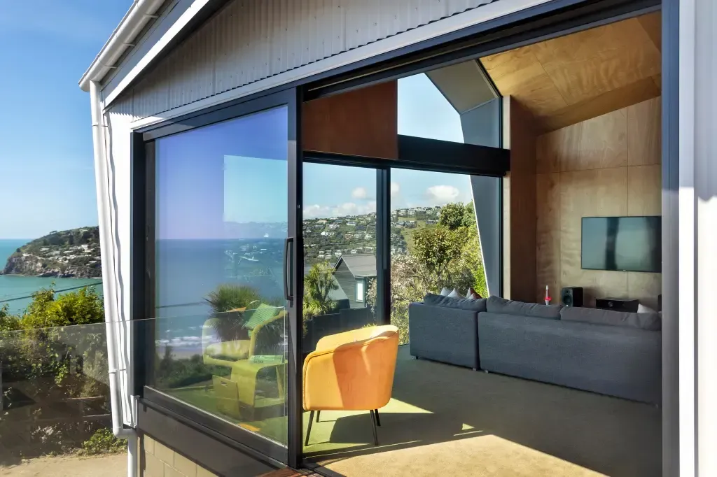 Modern house with open glass doors overlooking a coastal view; orange chair and gray sofa inside