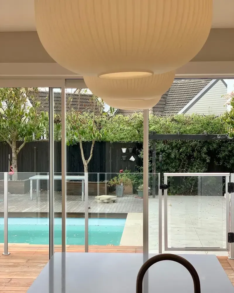 Indoor view of a backyard pool and patio through glass doors, with hanging lamps overhead