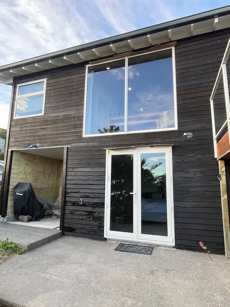 Two-story dark wood house with large windows, glass double doors, and a covered carport.