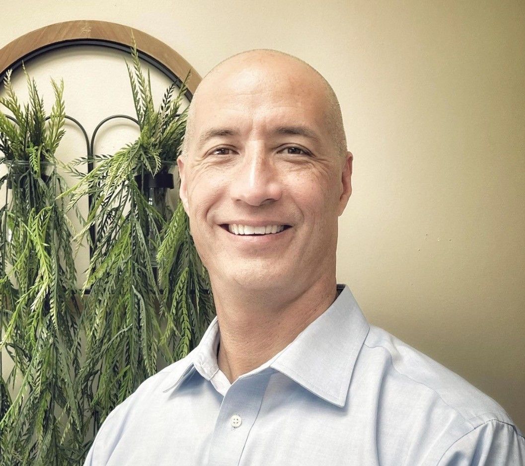 Smiling bald man in a light shirt standing indoors beside hanging green plants.