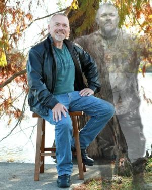 Man seated on a wooden stool beside a translucent ghostly figure outdoors under autumn leaves