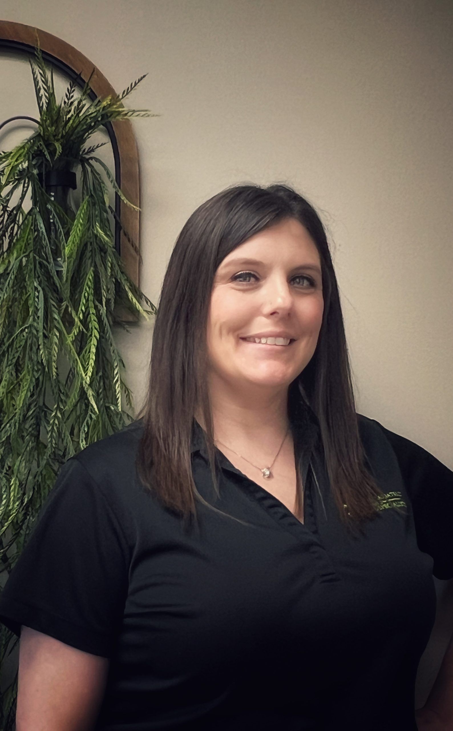 Smiling woman in a black shirt standing indoors beside a hanging plant.