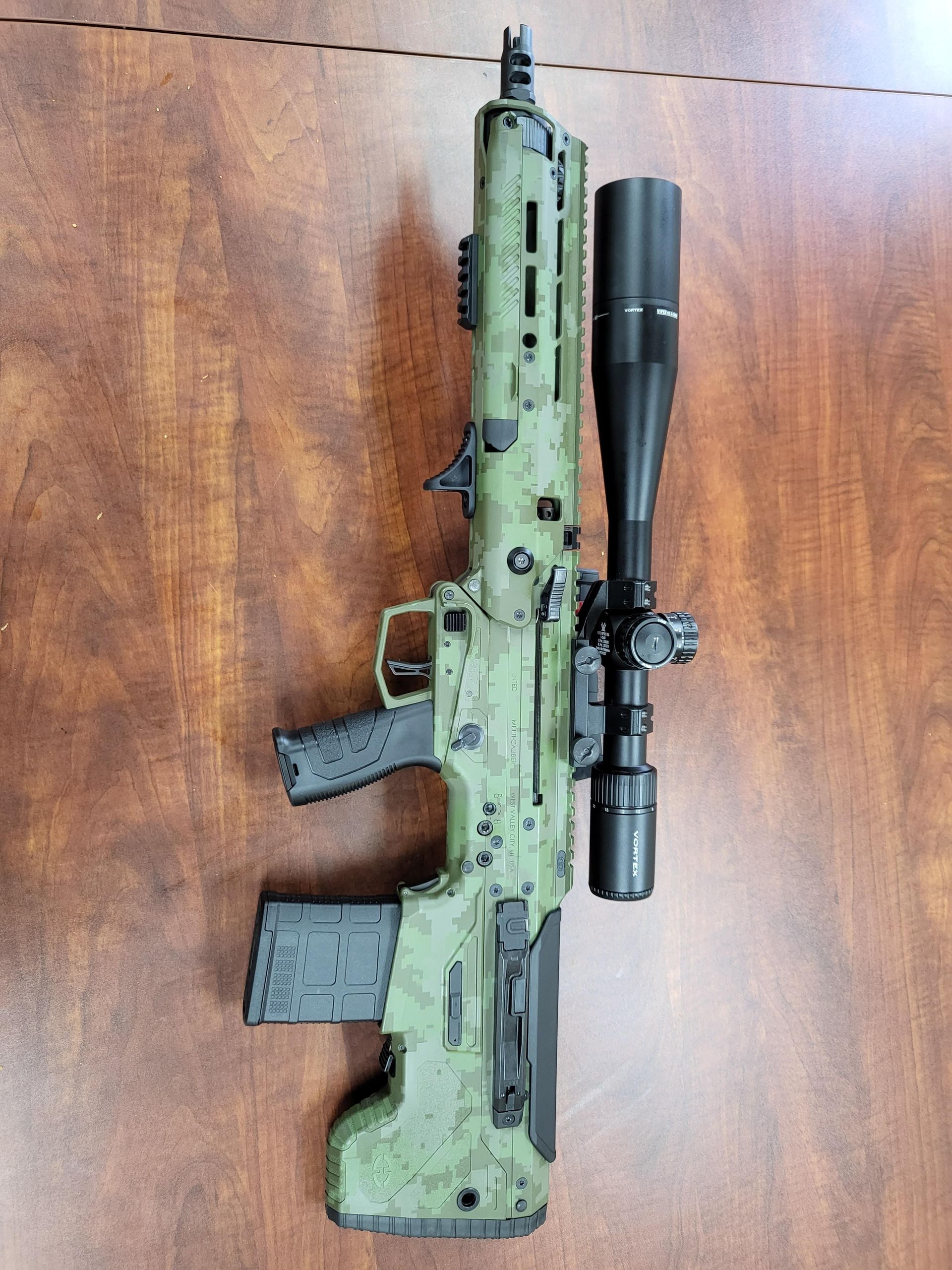 Green camouflage rifle with mounted scope and magazine lying on a wooden surface.