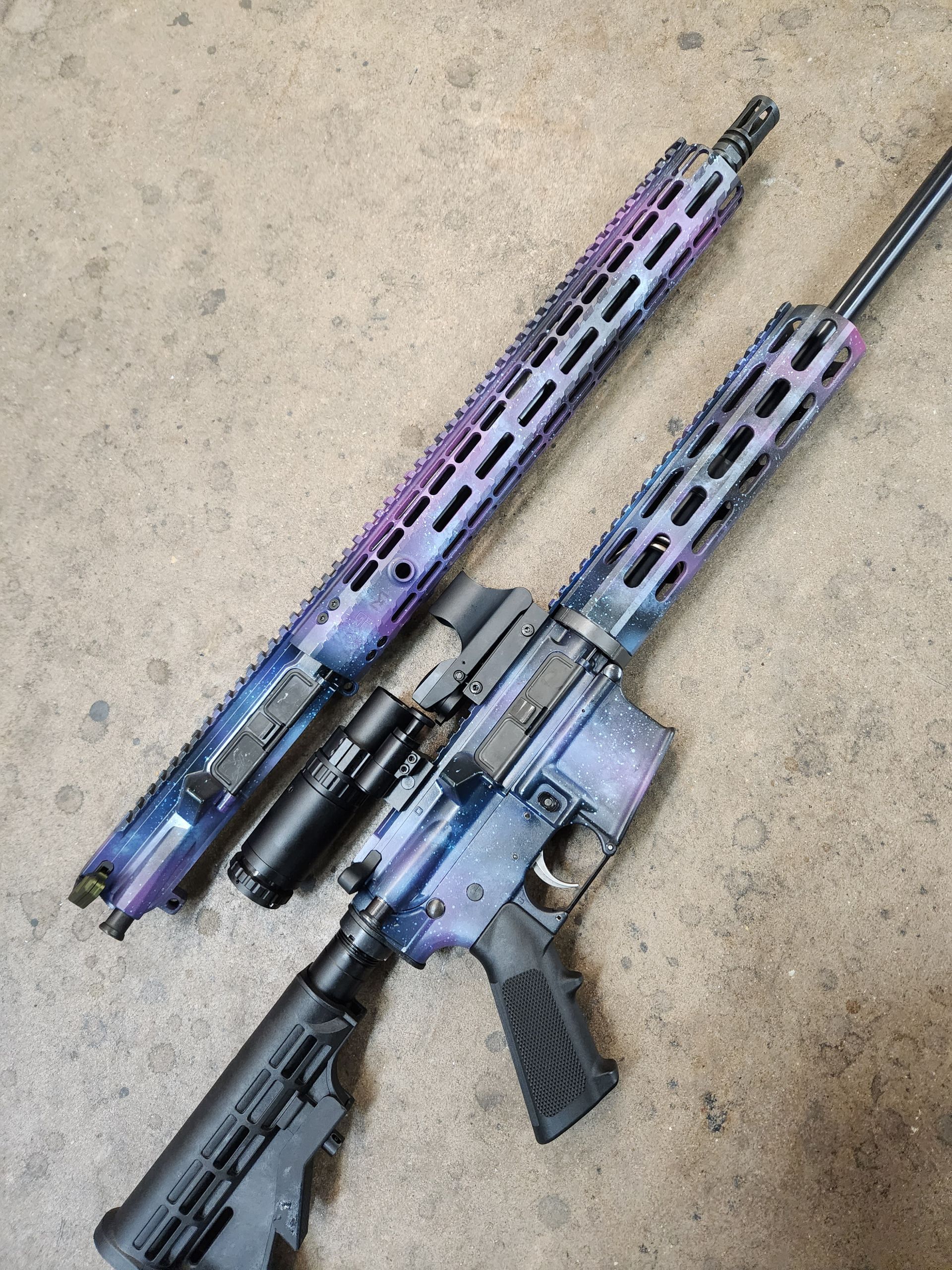 Two custom-painted rifles and a handgun laid on concrete, with purple-blue camo and black grips