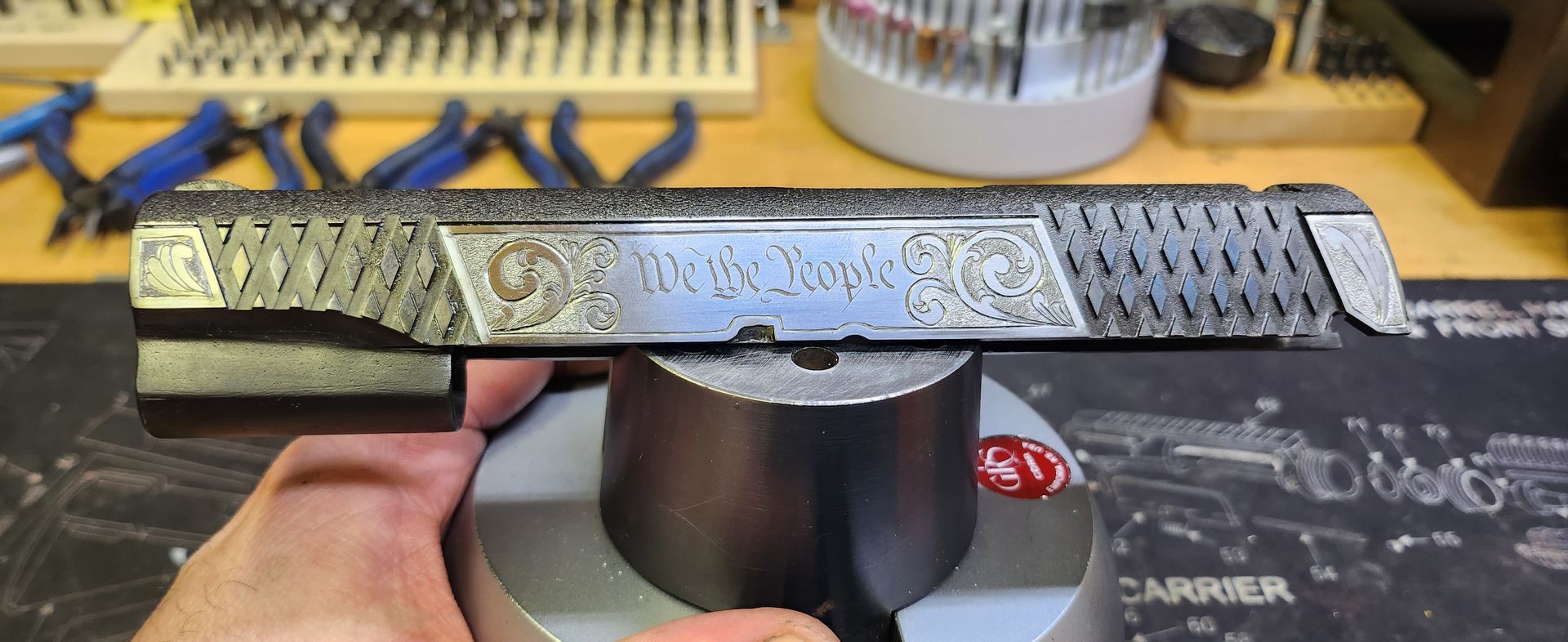 Hand holding a patterned metal handgun-shaped object on a workbench with tools in the background