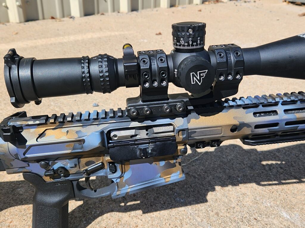 Scoped rifle resting outdoors on a concrete surface against a beige wall.