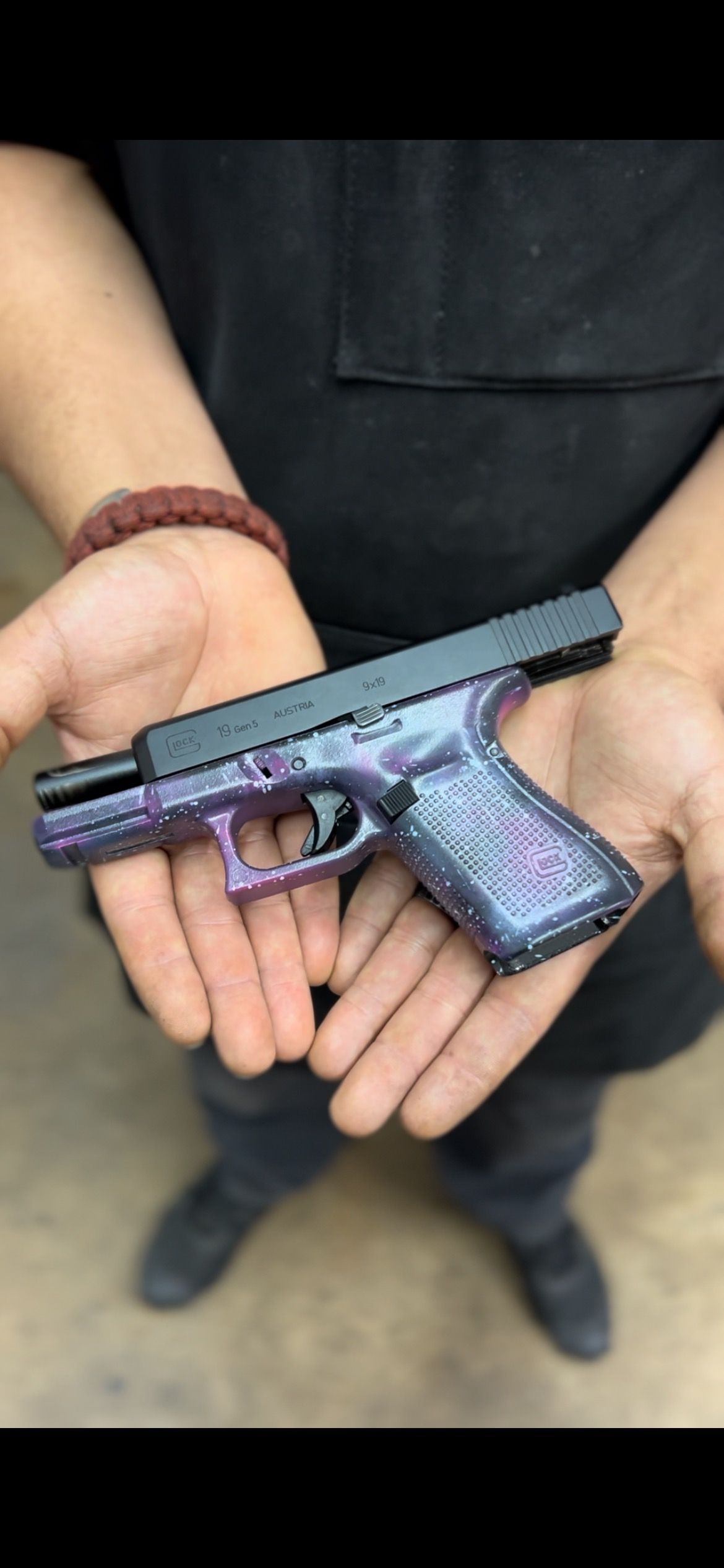 Person holding a purple-and-black handgun over a beige floor, wearing a black shirt and beaded bracelet