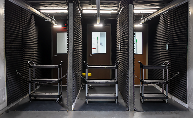 Three soundproof booths with black acoustic foam walls, each containing a rolling cart, inside a building interior.