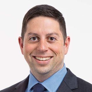 Professional headshot of a smiling person with short dark hair, wearing a dark suit and light blue button-down shirt.