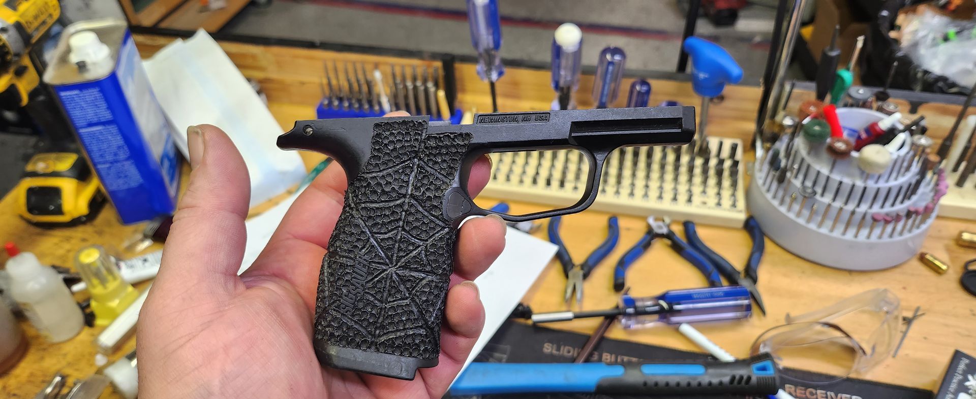 Hand holding a black textured pistol-shaped tool over a cluttered workbench with tools and parts